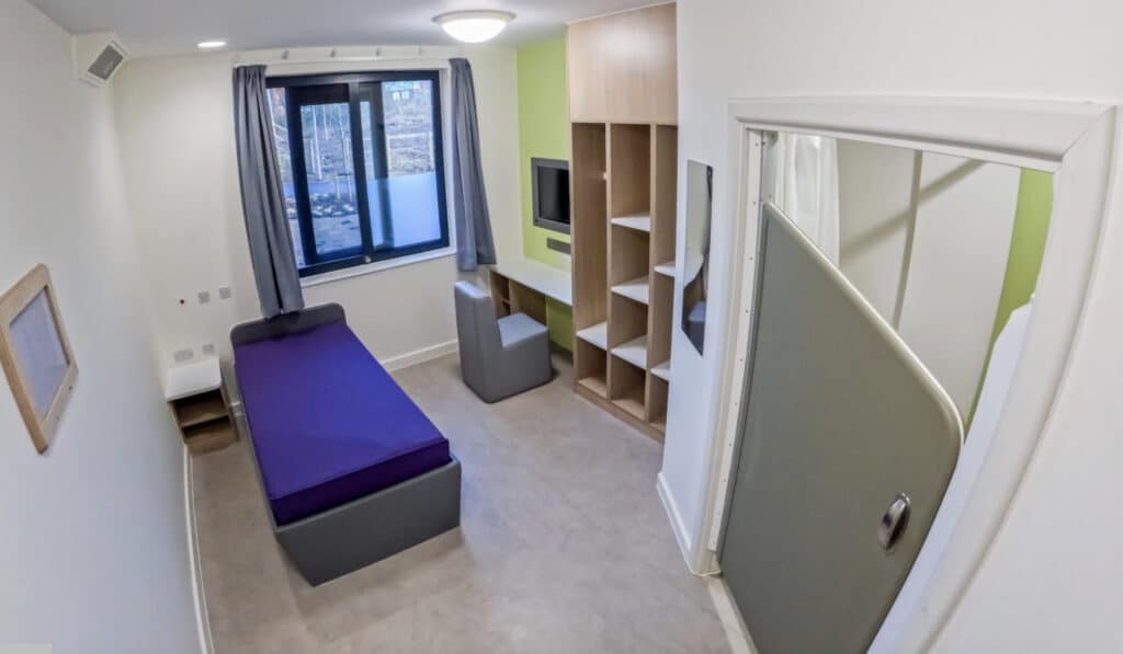 Healthcare room with small single bed, built-TV and desk, plus shelving. The room is light with a window looking out to new planted trees.