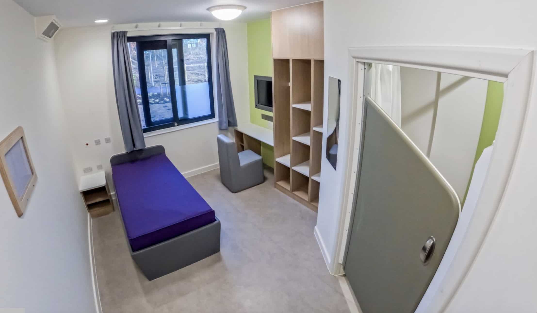 Healthcare room with small single bed, built-TV and desk, plus shelving. The room is light with a window looking out to new planted trees.