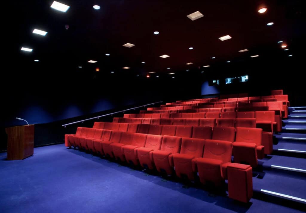 Dark theatre room with rows of red velvet covered chairs. staggered on different levels, directed towards a large stage area. A vibrant purple carpet lines the floor.