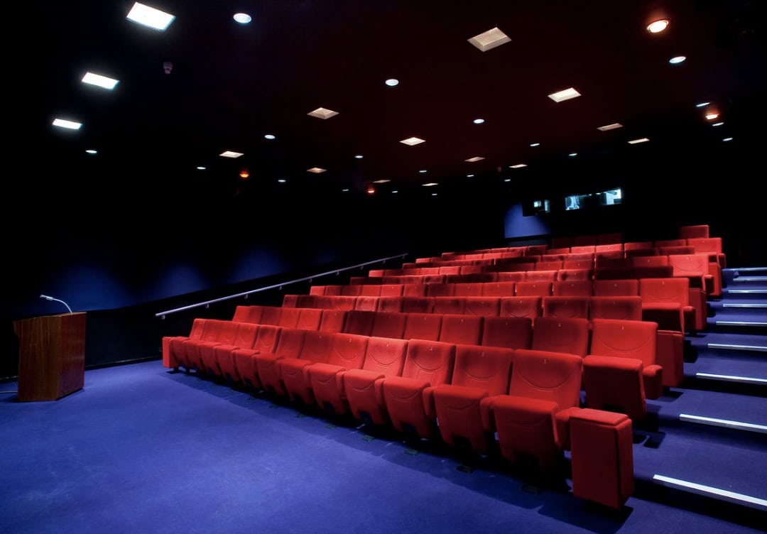 Dark theatre room with rows of red velvet covered chairs. staggered on different levels, directed towards a large stage area. A vibrant purple carpet lines the floor.