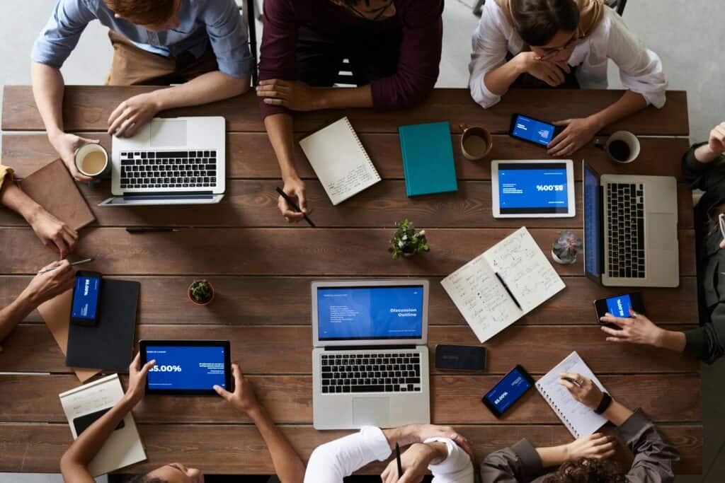 Image of office worker sat around a large wooden tables with laptops and note pads.